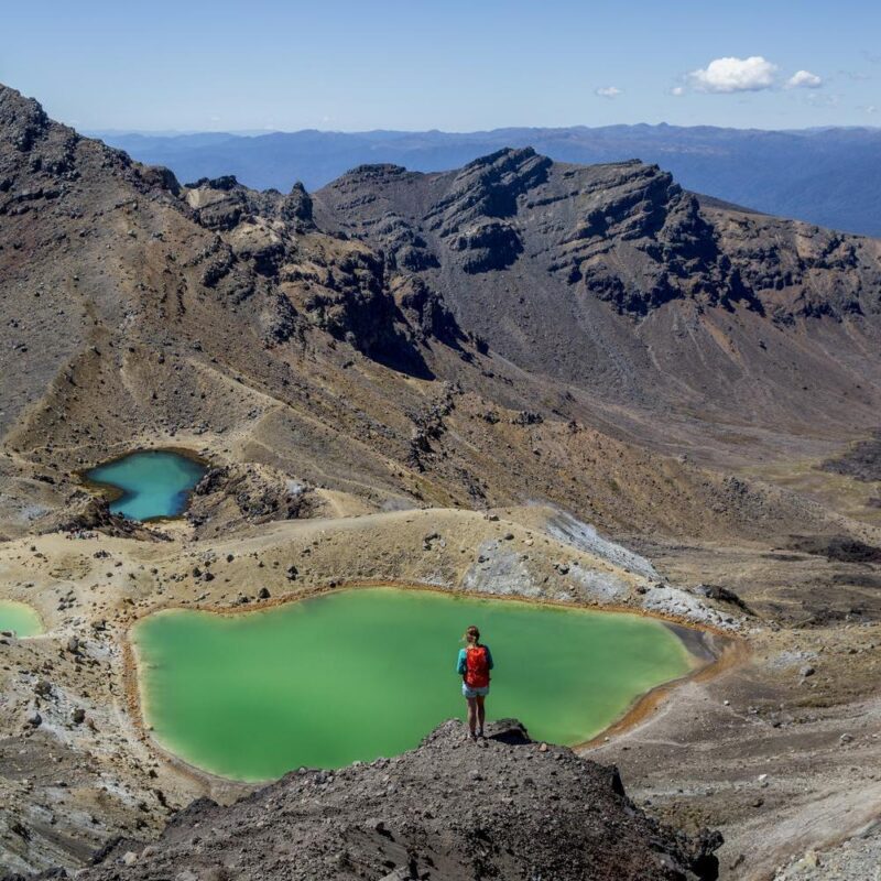 A woman stands looking over a bright green crater lake and volcanic landscapes in Tongariro, New Zealand
