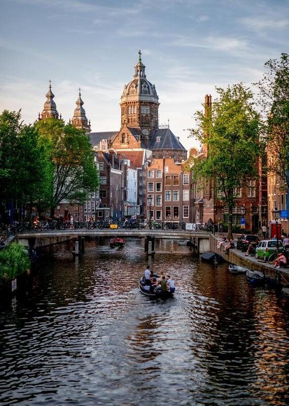 A small boat navigates an Amsterdam canal towards a brick bridge. The Basilica sits beyond.