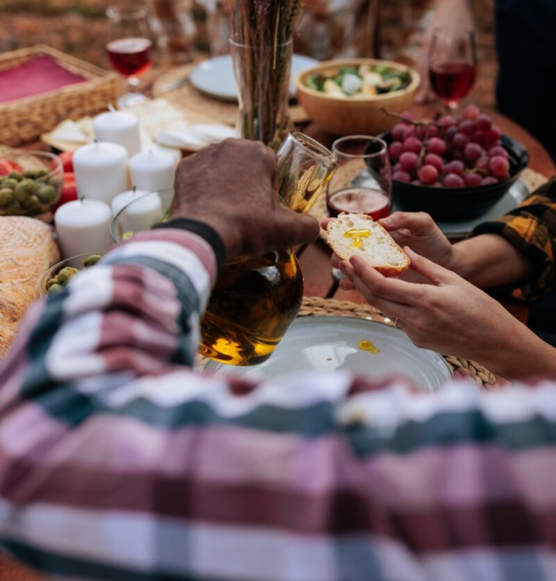 Friends are enjoying time together, pouring olive oil on bread during a festive outdoor picnic with wine and various food
