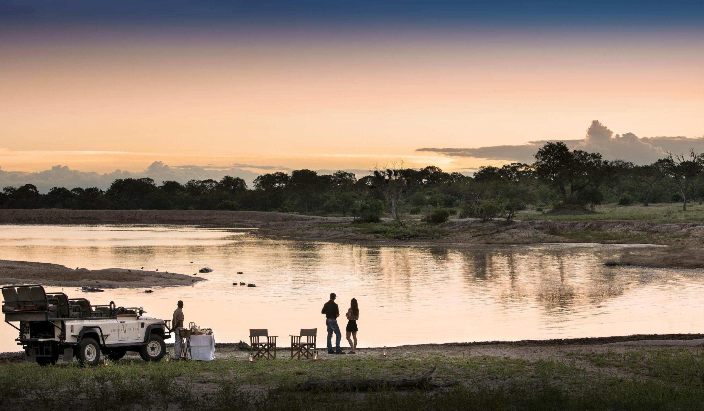 A couple standing by a river next to a safari vehicle at sunset during luxury Africa honeymoons.