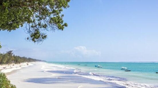 Lush green trees overlooking a long, white sand beach with gentle waves and small boats on a turquoise sea.