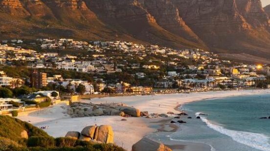 A scenic view of a coastal city nestled between steep mountains and a sandy beach with large rocks.