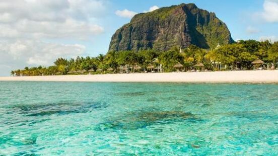 Eye-level view of a clear turquoise ocean with a sandy beach, tropical trees, and a large mountain in the background.