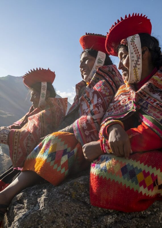 Women in colorful traditional hats and patterned clothing sitting outdoors to show positive impact.