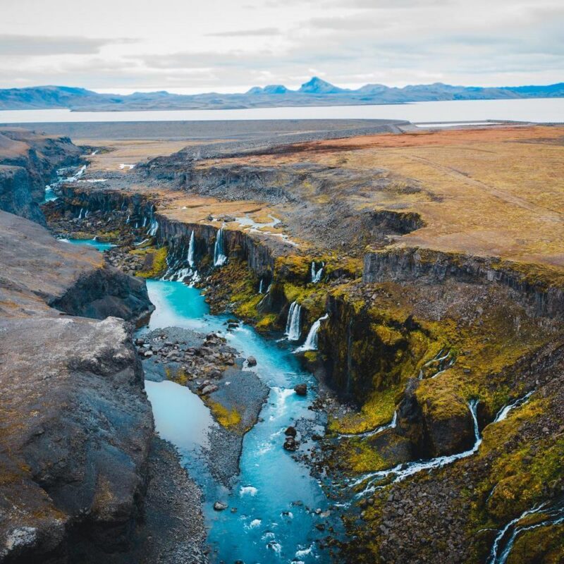 Aerial view of a bright blue river flowing through a rocky canyon with multiple waterfalls in Iceland.