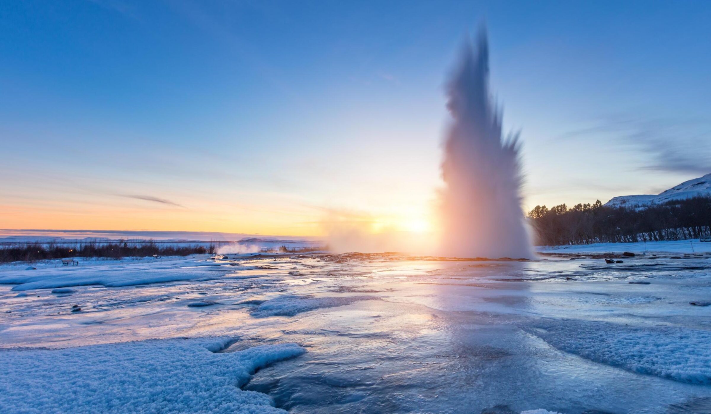 A high plume of water erupts from a geyser in a snowy field at sunset under a clear blue sky.