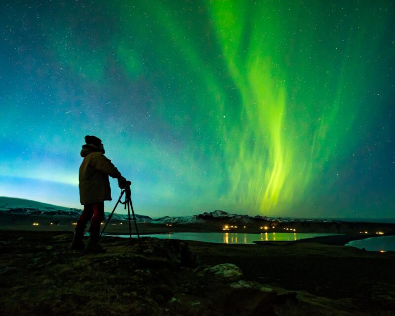 Bright green northern lights dancing in a starry sky over a silhouette of a photographer with a tripod.