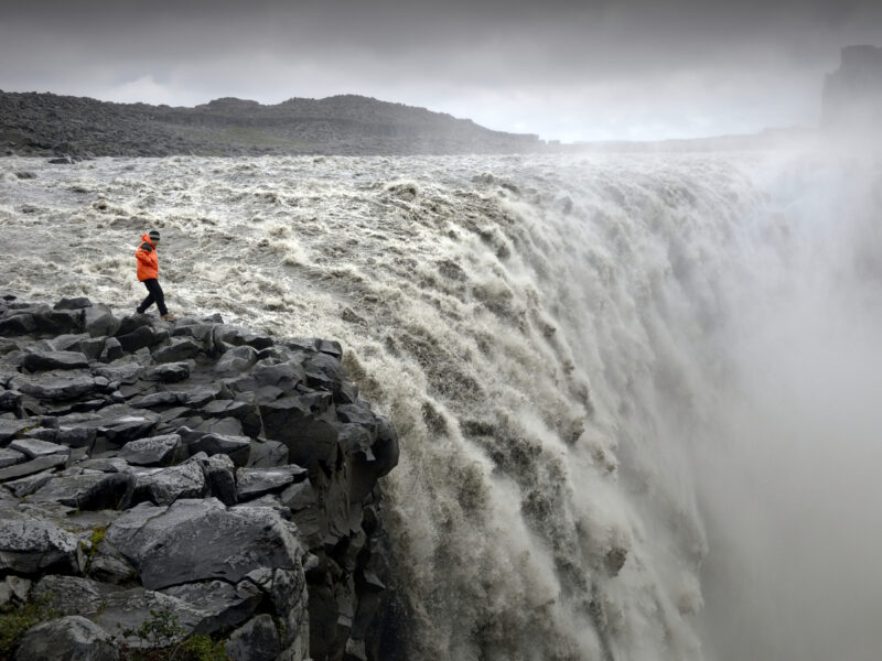 Large, powerful waterfall flowing over rocky cliffs with a person in an orange jacket standing on the edge.