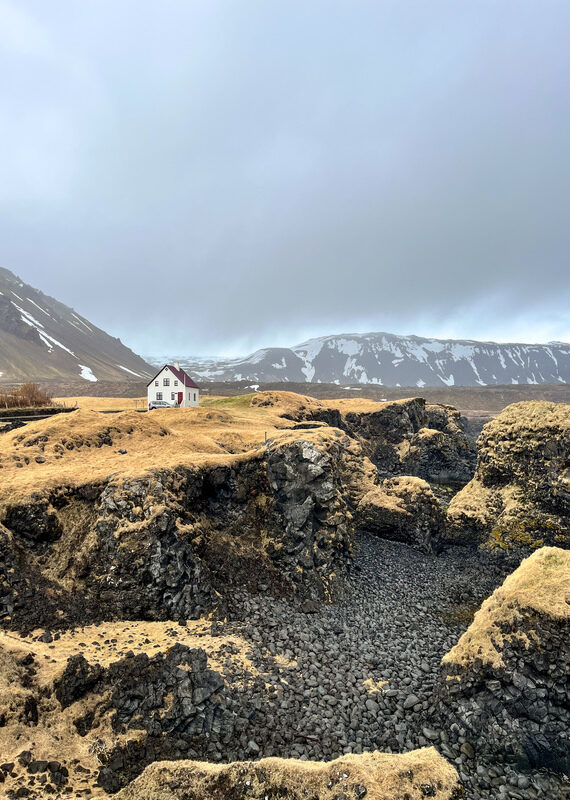A white house with a red roof on a grassy cliff overlooking dark, rocky terrain and snowy mountains.