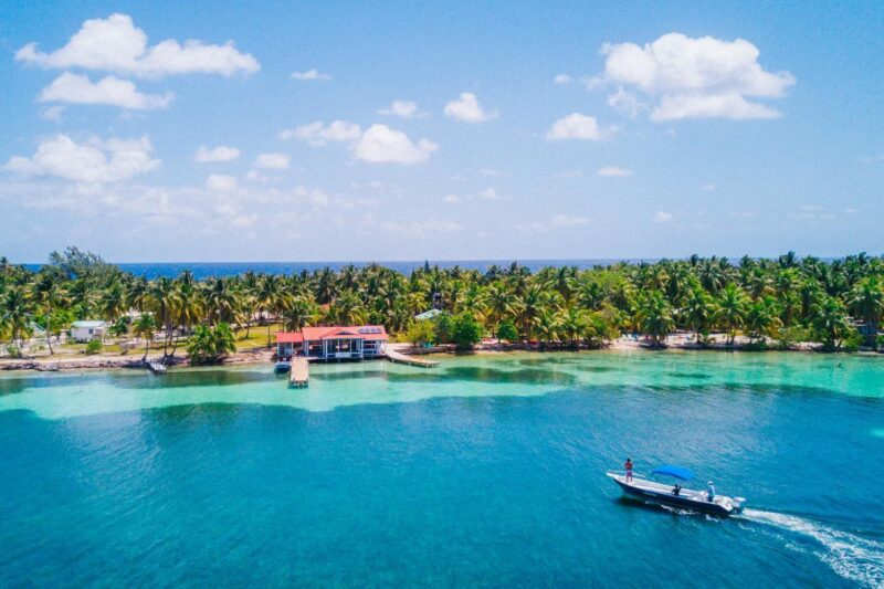 Aerial view of a Belize island with palm trees, a red-roofed building, and a boat on turquoise water.
