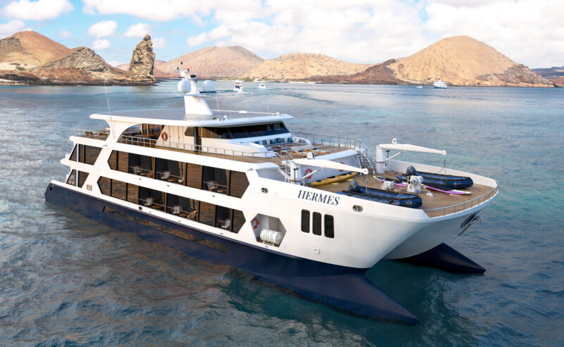 A large white and blue luxury catamaran named Hermes sailing on the ocean with volcanic hills in the background.