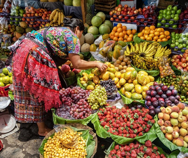 Woman wearing colorful traditional attire arranging mangoes at a vibrant outdoor market with strawberries, grapes, and bananas.