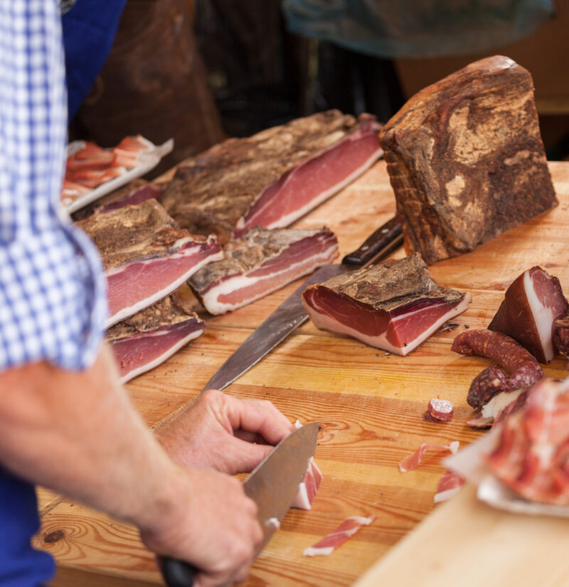 Close-up of hands slicing smoked cured meat on a wooden table at an outdoor market.