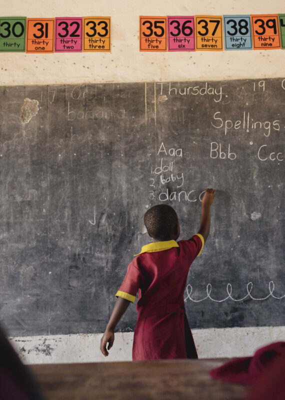 A young child with her back to the camera, writes on a classroom chalkboard.