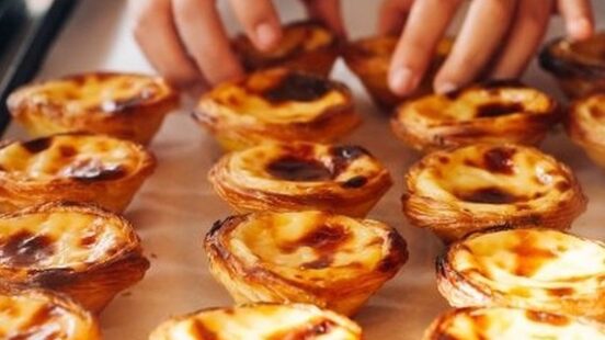 A person in a white chefx27;s coat arranges a tray of freshly baked custard tarts with caramelized tops.