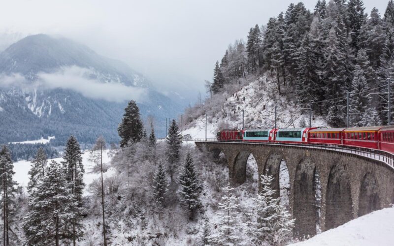 A red and white train crosses a stone viaduct over a snowy forest in winter, a beautiful sight on luxury Europe rail tours.