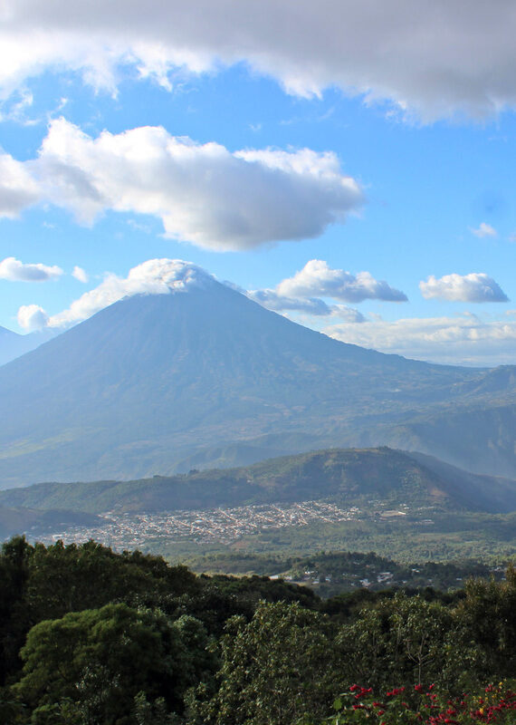 A large volcano partially covered by clouds towers over a valley and town in the Guatemalan Highlands.
