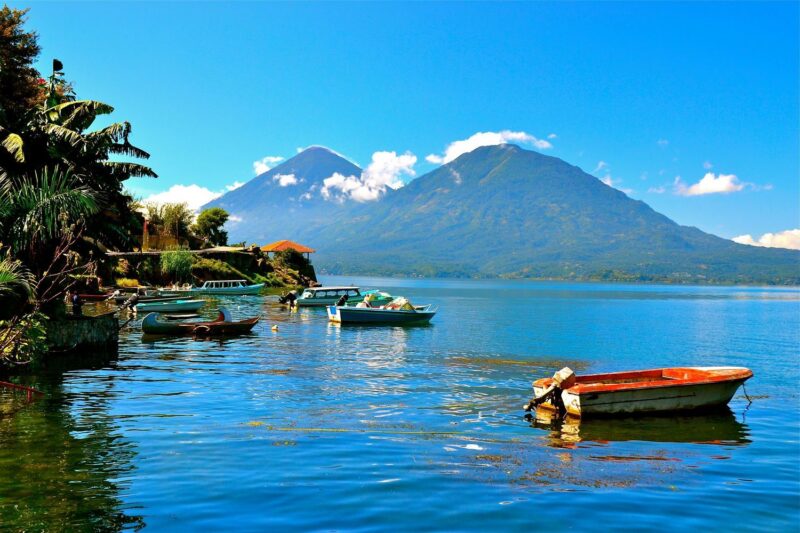 Several small boats docked on a vibrant blue lake with large volcanoes and green foliage in the background.