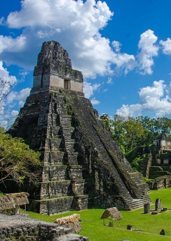 A tall ancient Mayan stone temple with steep steps stands against a bright blue sky with white clouds.