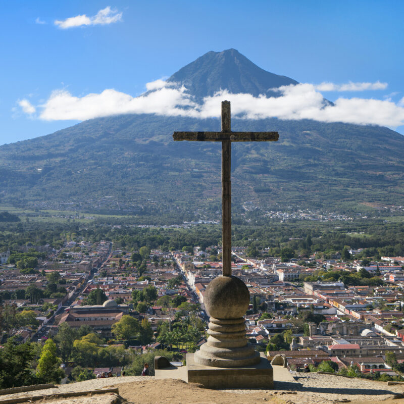 A large stone cross on a pedestal overlooks a city valley toward a massive volcano under a blue sky.