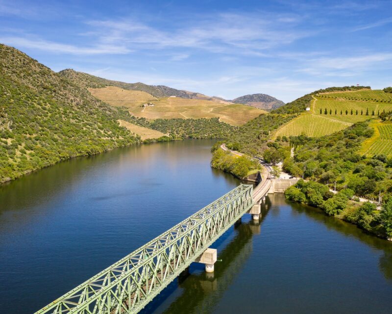 An aerial view of a green metal railway bridge crossing a wide river, with vineyard-covered hills, part of many luxury Europe rail tours.