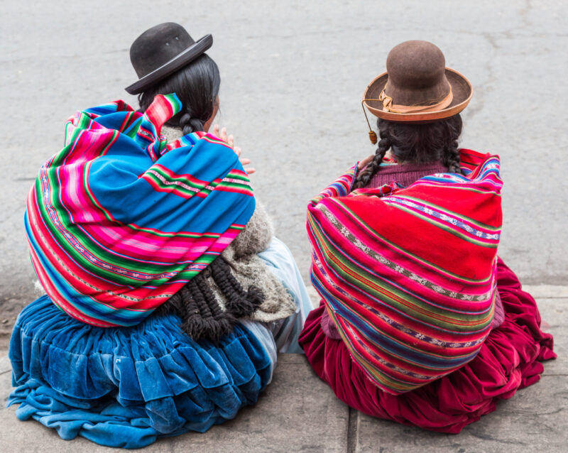 Two women with braids wearing traditional clothes sit on the streets of Puno city in Peru