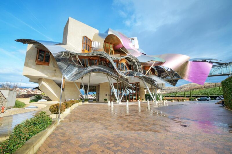Modern building with flowing pink and silver titanium panels over a stone structure under a bright blue sky.