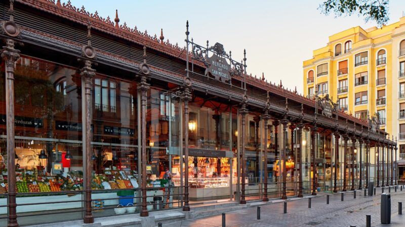 Exterior view of a glass-walled market hall with ornate dark iron pillars and a sign reading Mercado de San Miguel.