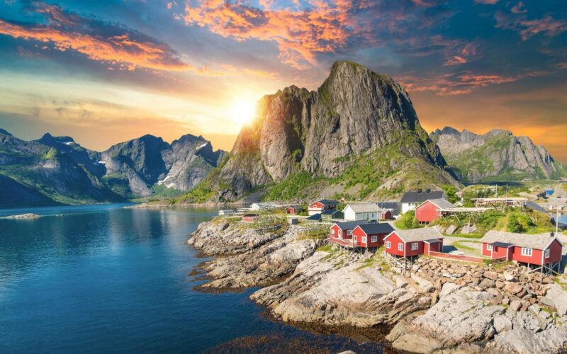 Evening sunlight over the red fishing cabins of the Lofoten Islands, with rugged peaks rising above the fjord in northern Norway.