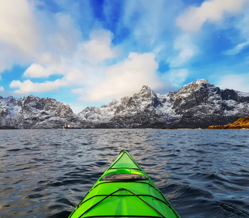 Small green kayak in bay fjord in the Lofoten Islands surrounded with snowy mountains and blue sky. Norway