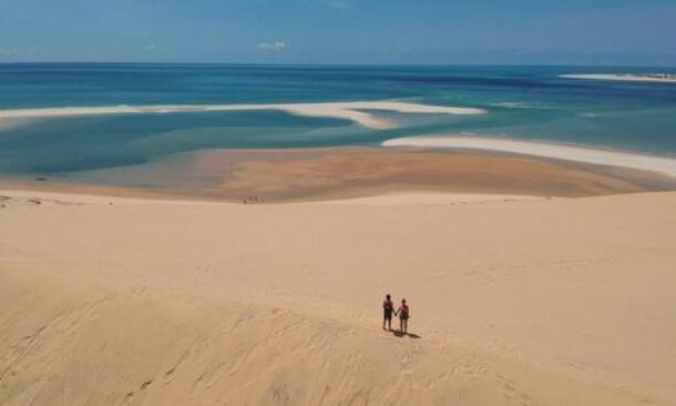 Two people standing on a vast tan sand dune with a view of turquoise water and sandbars in the distance.