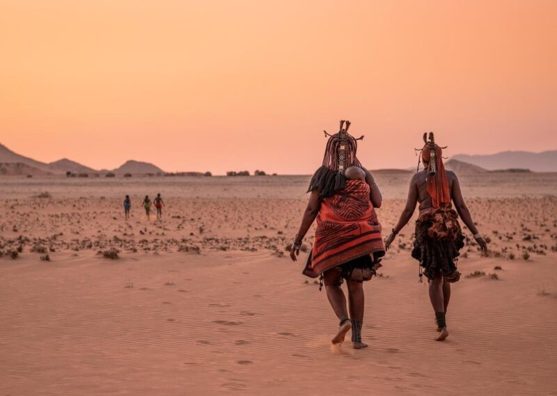 A group of children play in the foreground while a Himba woman with a young child wrapped onto her back walks beside another Himba woman.