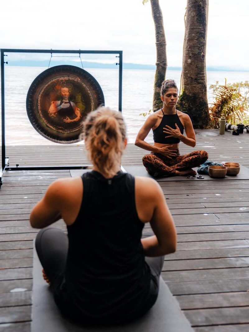 Two women in a meditative seated pose on a wooden platform with the ocean behind them