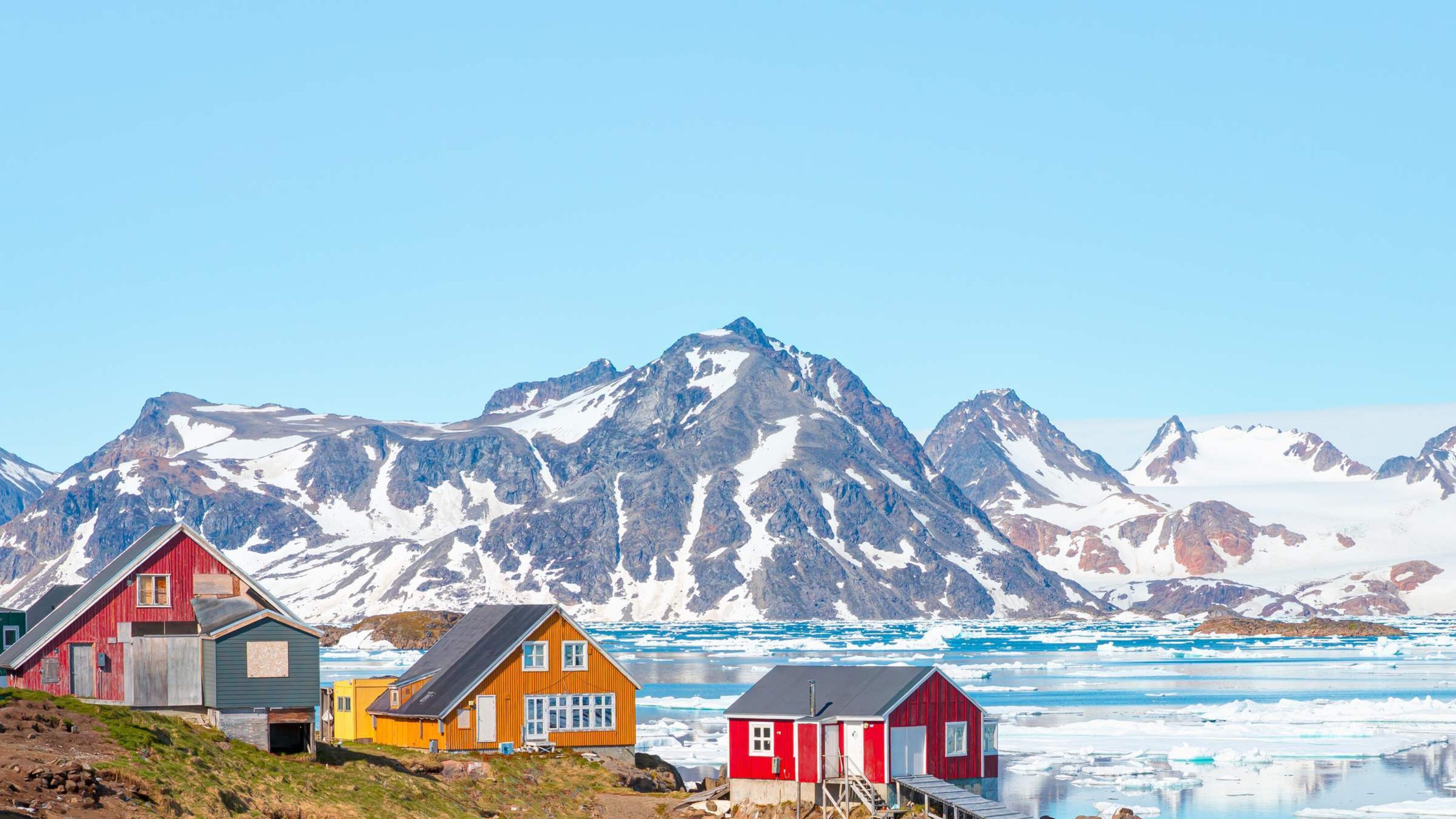 Three colourful houses built next to the water with mountains in the background in Greenland