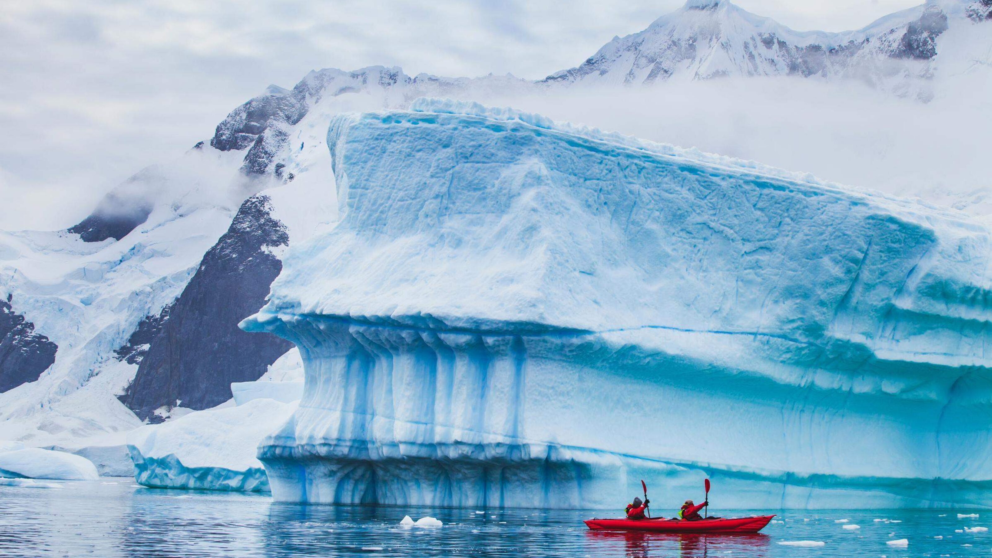 Two people in a kayak paddling in front of a giant iceberg.