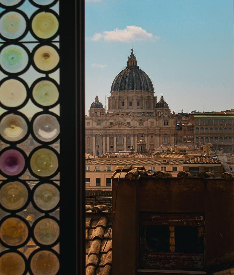 View through an open stained glass window over rooftops and St Peter's Basilica in Rome.