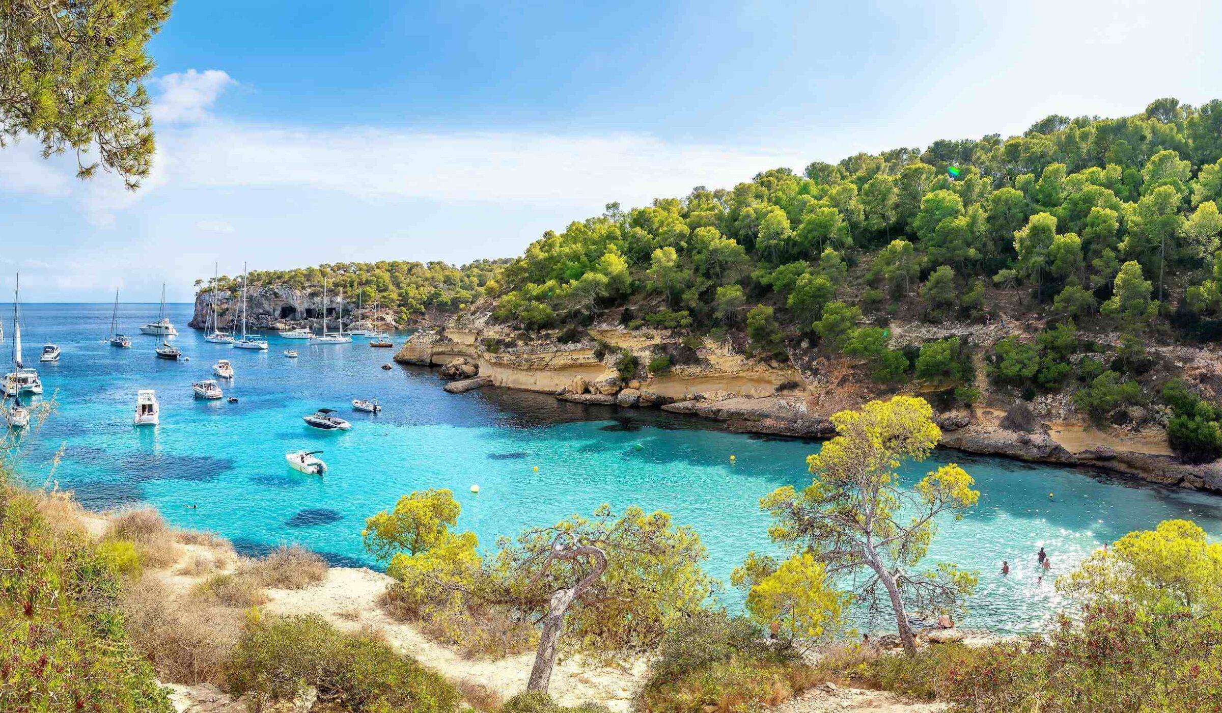 View of a bay with boats floating in turquoise water, surrounded by rocks and trees in Mallorca, Spain