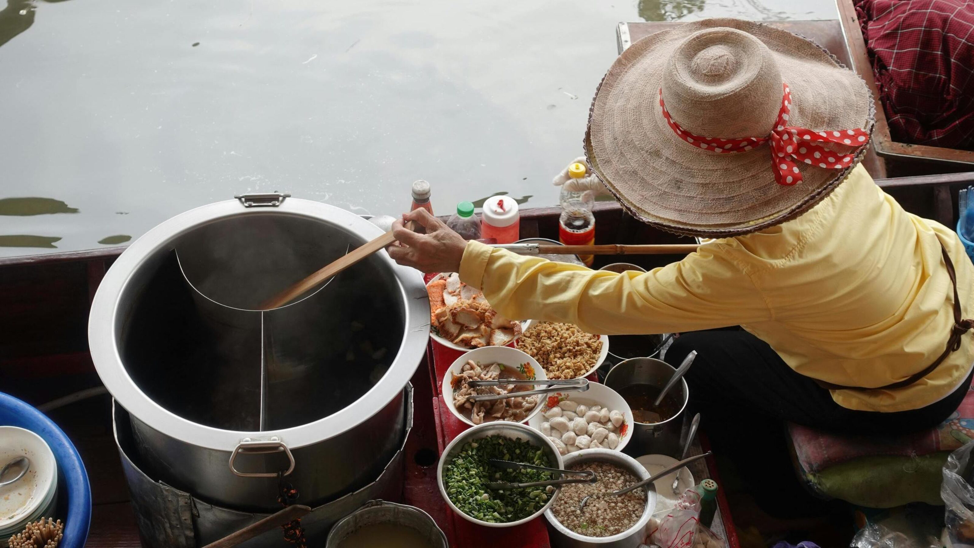 Top-down view of a floating market vendor preparing food in a large pot on a wooden boat.