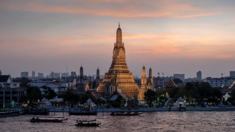 Illuminated Wat Arun temple at sunset overlooking the Chao Phraya River in Bangkok.