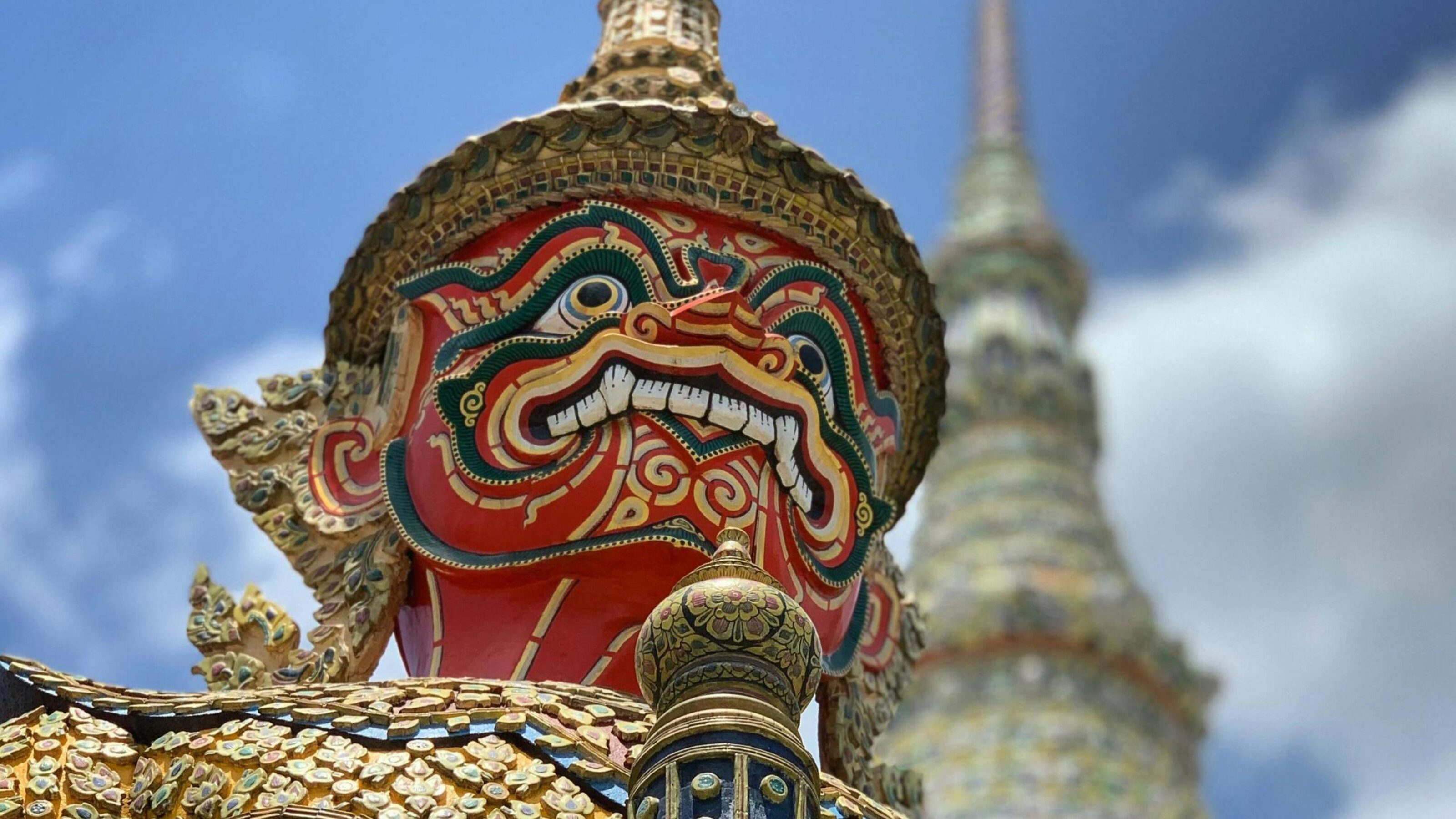Close-up of a red and gold ornate Thai temple guardian sculpture against a blue sky.