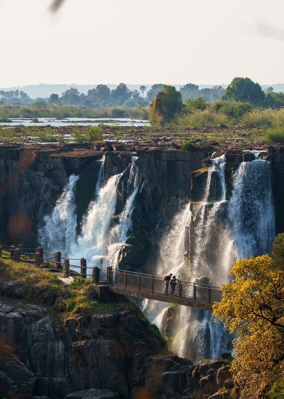 Two people on a walkway bridge look out over the rushing white water of Victoria Falls.