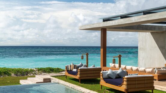 Luxury resort deck with cushioned outdoor lounges and a stone pool facing turquoise ocean waves under a bright sky.