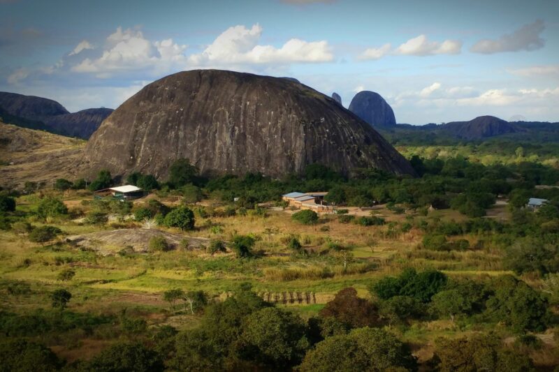 A giant dome-shaped rock formation rises above a rural landscape with scattered trees and small structures.
