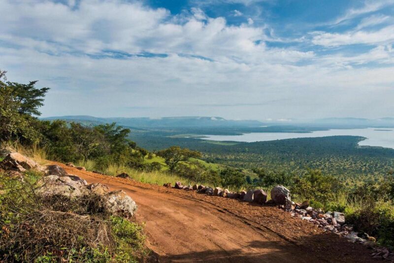 A red dirt road curves along a grassy hillside overlooking a vast lake and distant mountains under a blue sky.