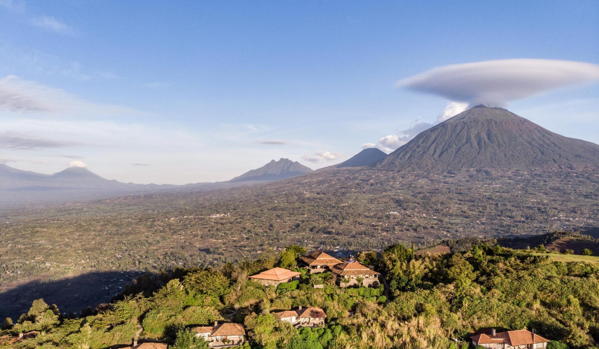 Aerial view of a luxury lodge on a lush green hilltop with large volcanoes in the background of Virunga National Park.