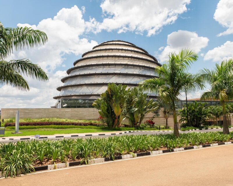 The modern, tiered dome of the Kigali Convention Centre behind tropical greenery and a bright sky.