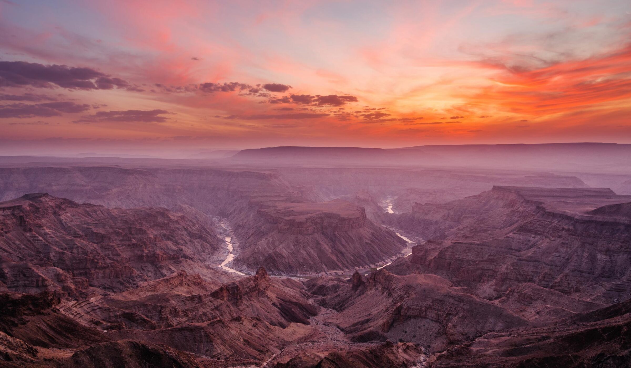 Panoramic view of a desert canyon landscape under a dramatic sunset sky with purple and orange clouds.