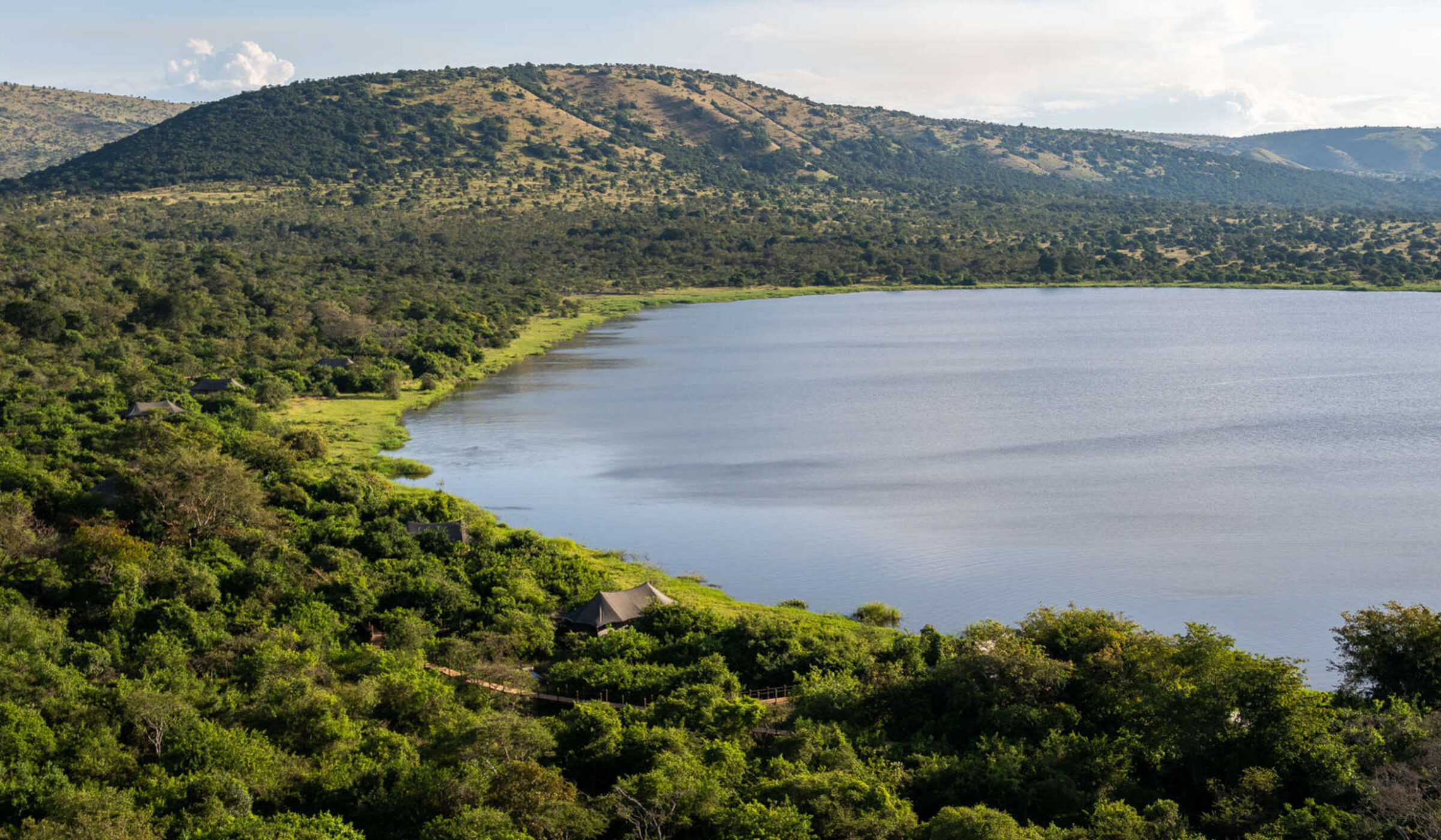 Wide view of a blue lake in Akagera National Park with lush green trees, safari tents, and hills under a clear sky.