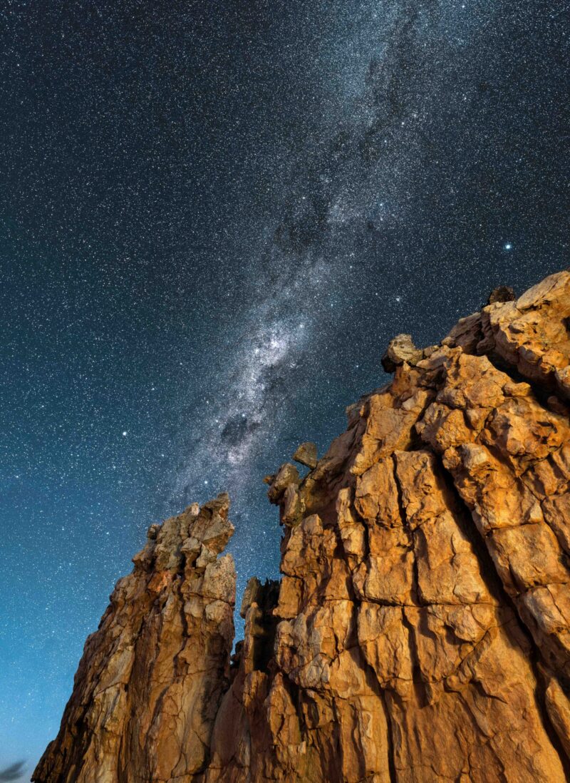 A beautiful vertical night sky landscape, with a yellow rock formation in the foreground and the Milky Way against a deep blue sky in the background, taken in the Cederberg mountains, South Africa.