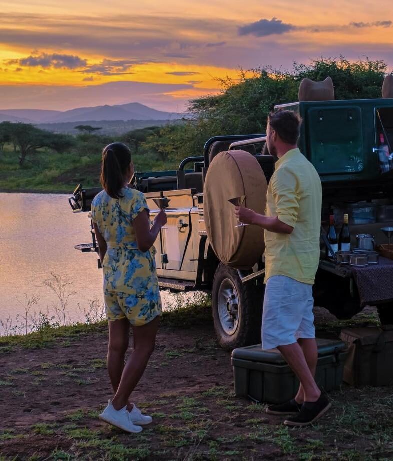 A couple stand by a lake at sunset enjoying sundowner drinks after a day on safari